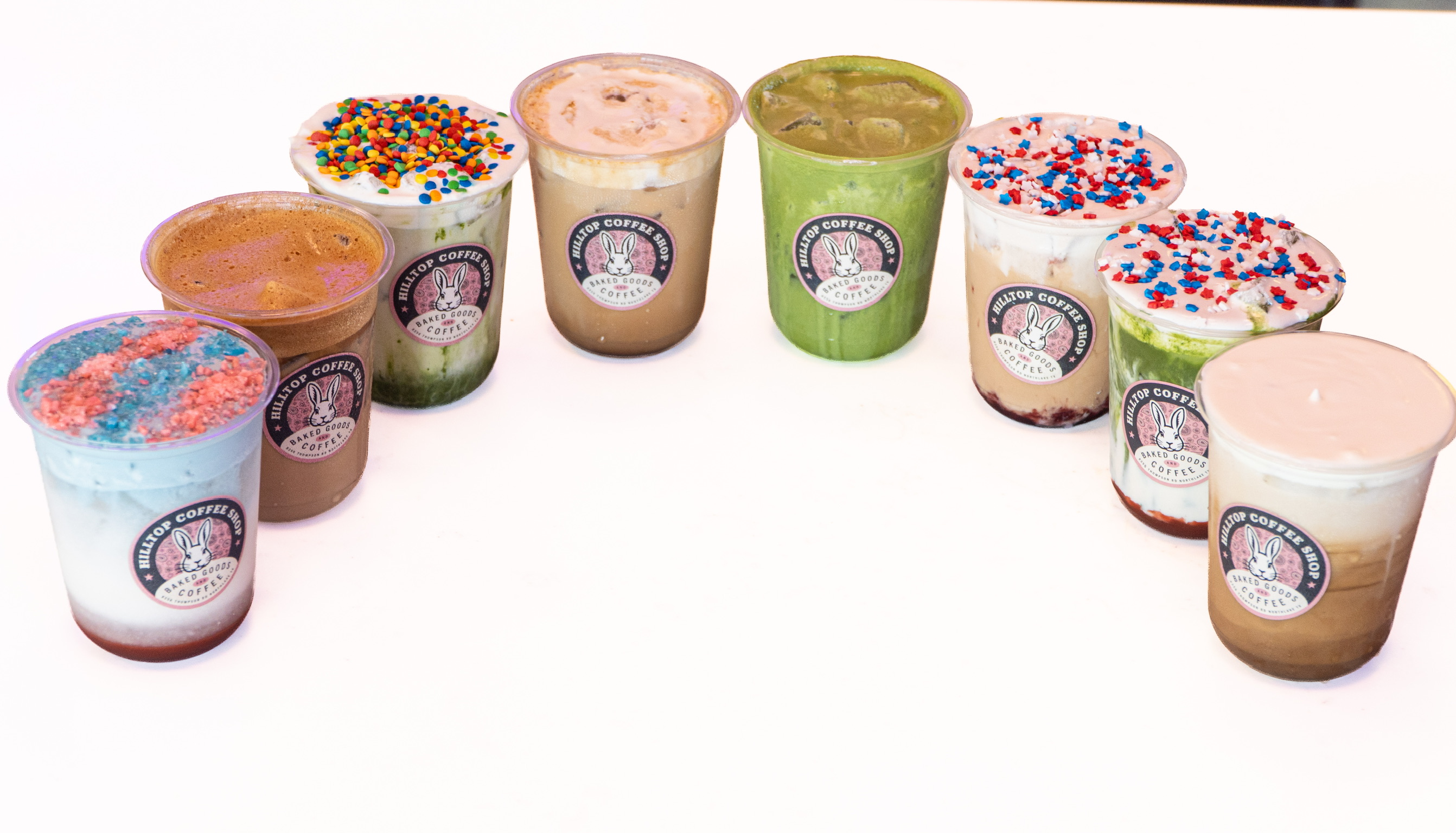 A lineup of Hilltop Coffee Shop July drinks arranged in an arc, each in a cup with the bunny logo, ranging from a red-white-and-blue refresher to matcha and cream-topped lattes